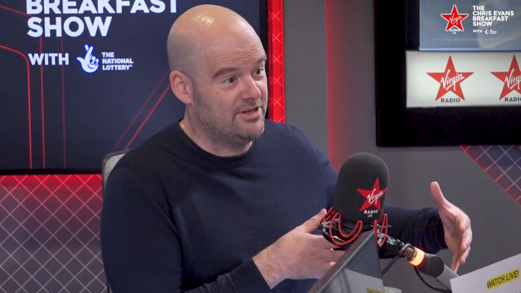 A man with a bald head and a short beard, wearing a dark blue crewneck sweater, speaks on the Virgin Radio UK breakfast show. He is sitting in a radio studio and gesturing with his hands toward a microphone with a red foam cover that has the Virgin Radio UK logo on it. This man is Dan Houser, a co-founder of Rockstar Games.