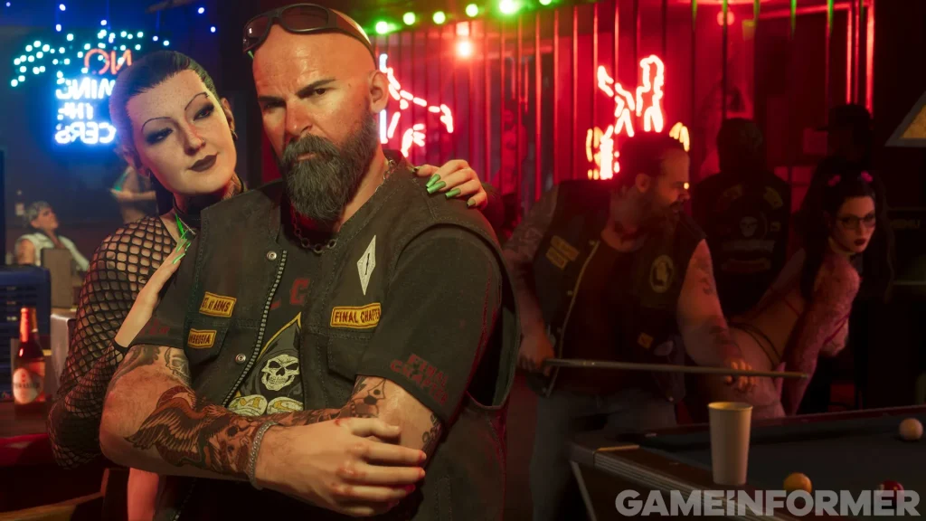 A Grand Theft Auto VI screenshot featured by Game Informer, showing a bald biker with a full beard and sunglasses folded on his head, wearing a leather vest with patches inside a neon-lit bar. A woman with dark hair and a mesh outfit leans over his shoulder, while other club members play billiards at a pool table in the background, with a Game Informer watermark in the bottom right corner.