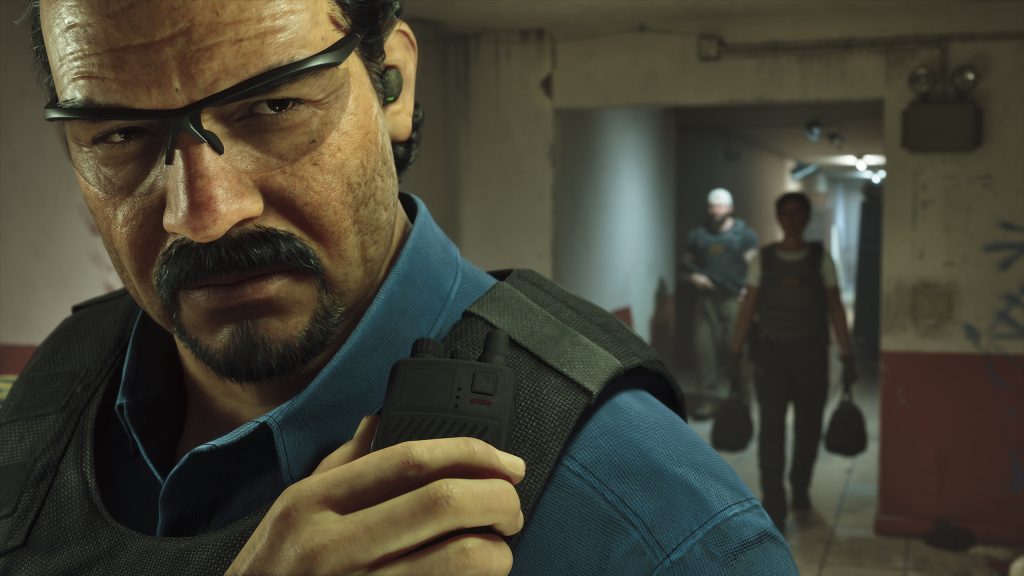 A close-up of Méndez, with a serious and intense look on his face, in a dim, urban environment. He wears a blue collared shirt and a tactical vest. In his right hand, he holds a small, black two-way radio to his shoulder. He has dark hair, a mustache, and a short beard. Large, black-framed shooting glasses are on his face. In the background, two individuals, indistinct and in shadows, stand in an open doorway. One has a white headcovering. The walls are cracked and show signs of wear.